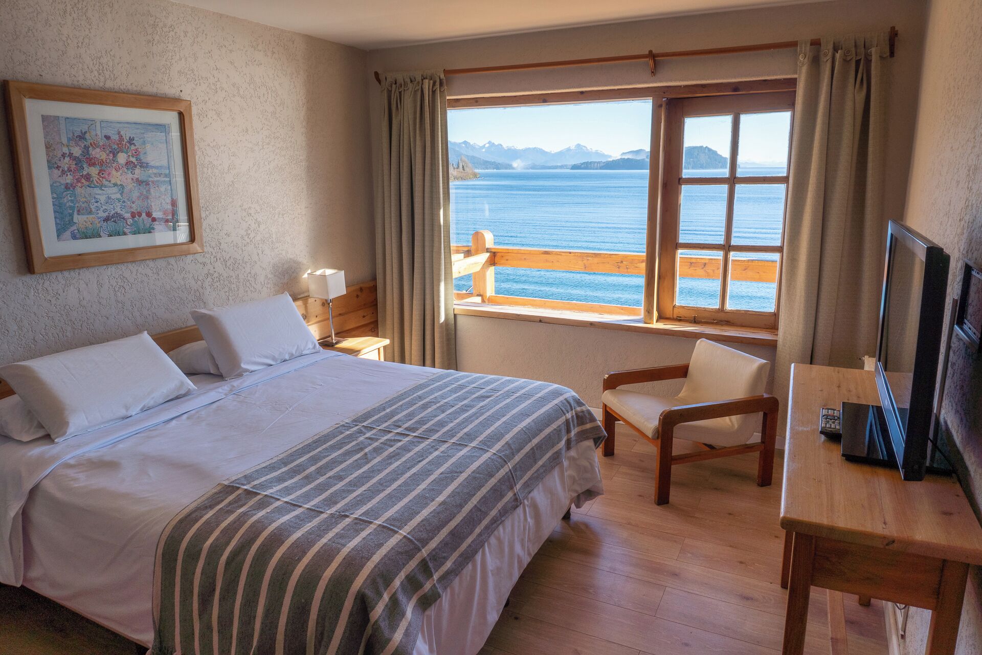 Une chambre d’hôtel avec un lit, une chaise, un bureau, une télévision, une fenêtre donnant sur la mer et un tableau au mur.