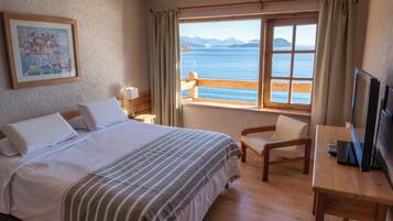 Une chambre d’hôtel avec un lit, une chaise, un bureau, une télévision, une fenêtre donnant sur la mer et un tableau au mur.