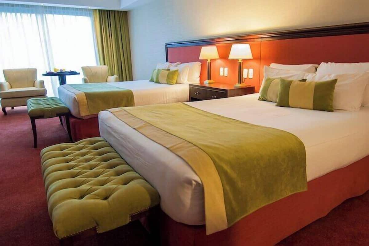 Une chambre d’hôtel avec deux lits, un coin salon comprenant une chaise et une petite table, et une grande fenêtre avec des rideaux.