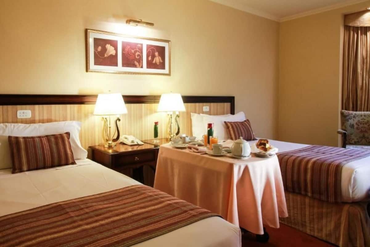 Une chambre d’hôtel avec deux lits, une table de chevet avec une lampe, une petite table avec une tasse et une assiette, et une chaise.