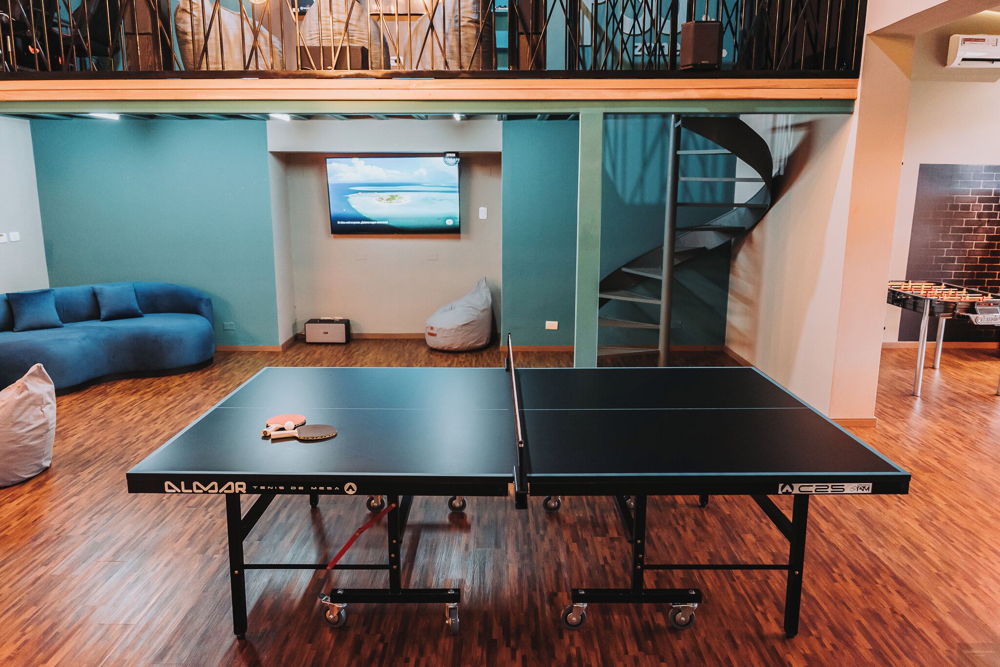 Una mesa de ping pong en una habitación con un sofá azul, un televisor y una escalera.