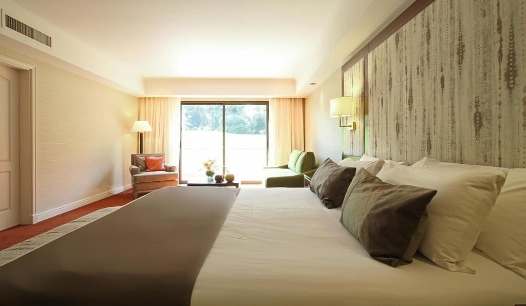 Une chambre d’hôtel dotée d’un grand lit, d’un coin salon avec un fauteuil et une table, d’une grande fenêtre et d’un mur au motif texturé.