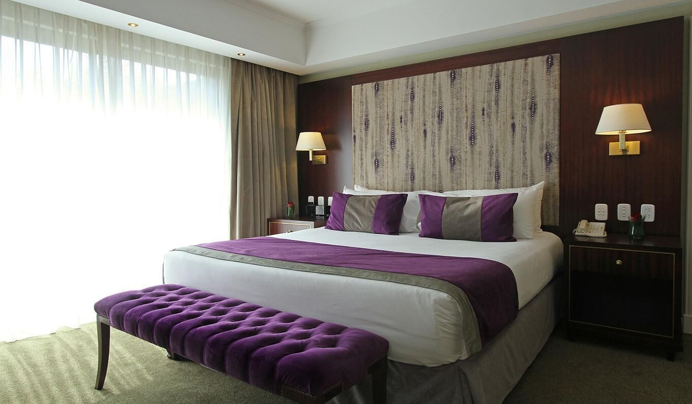 Habitación de hotel con una cama grande, dos lámparas de noche, una mesita de noche, un banco y vistas al exterior.