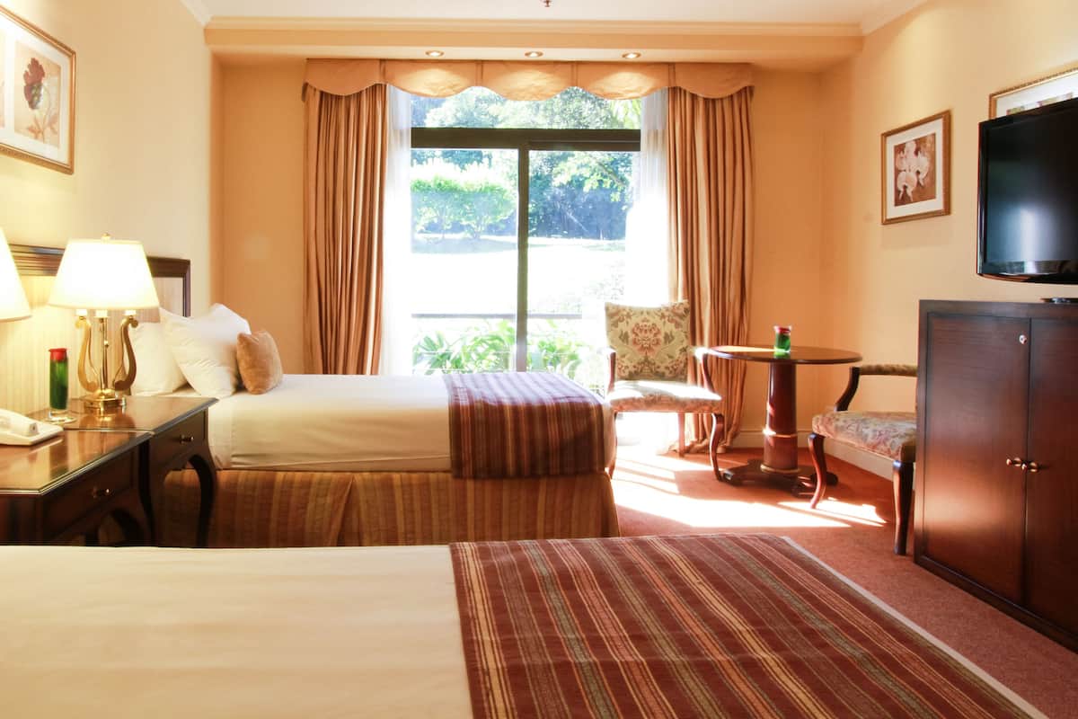 Une chambre d’hôtel avec deux lits, un bureau avec des lampes, une télévision et une vue sur la verdure environnante.