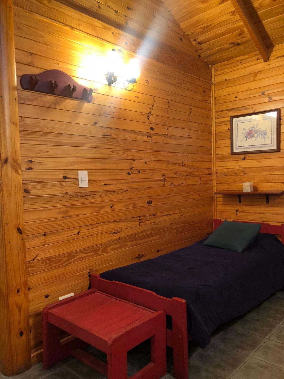A wooden cabin room with a bed, a small table, and a wall-mounted shelf with decorative items.