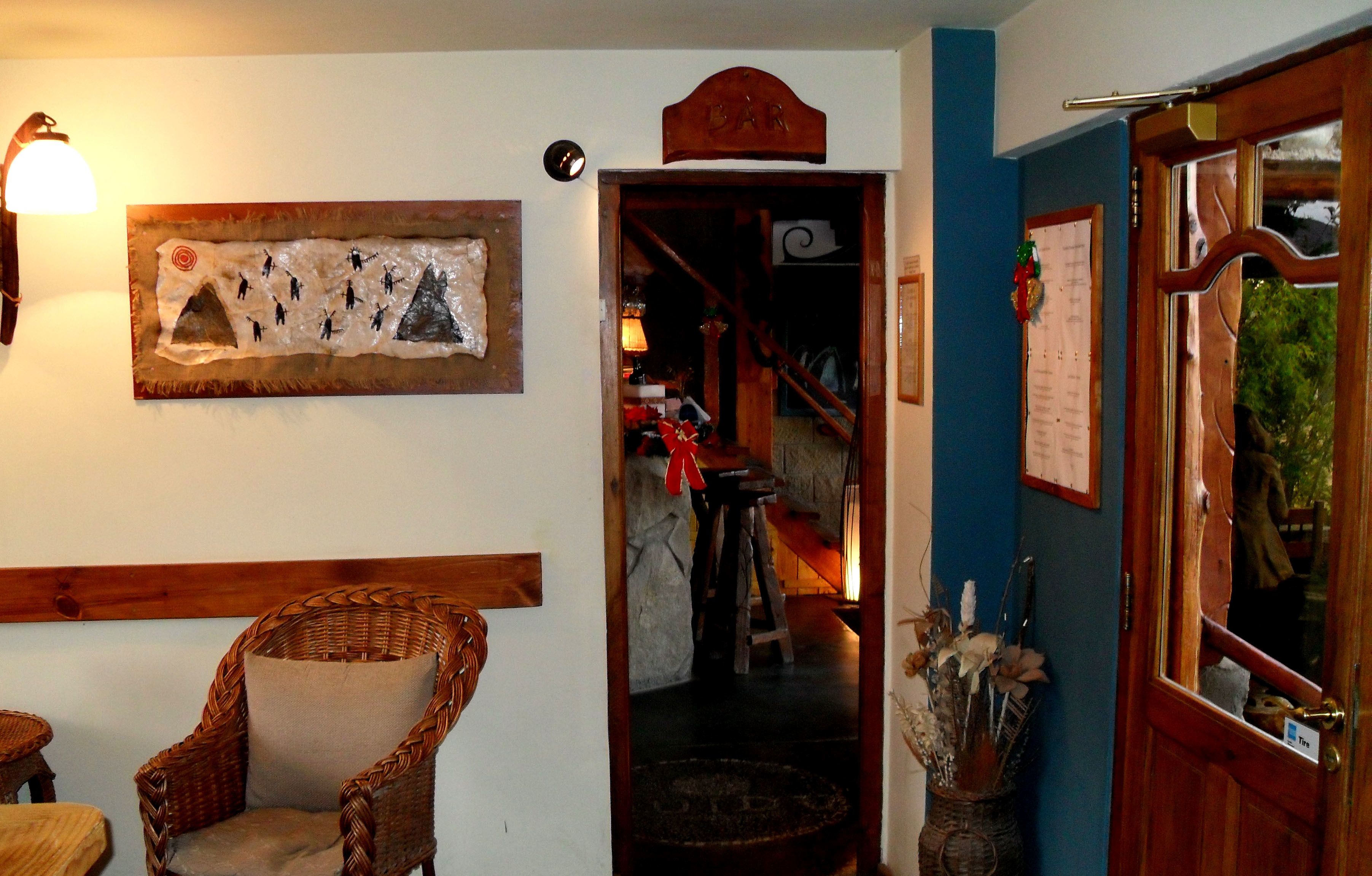 Una habitació amb una cadira de vimet, una obra d'art penjada a la paret, una porta de fusta amb un rètol que diu 'BAR' i una escala que porta a un nivell inferior.