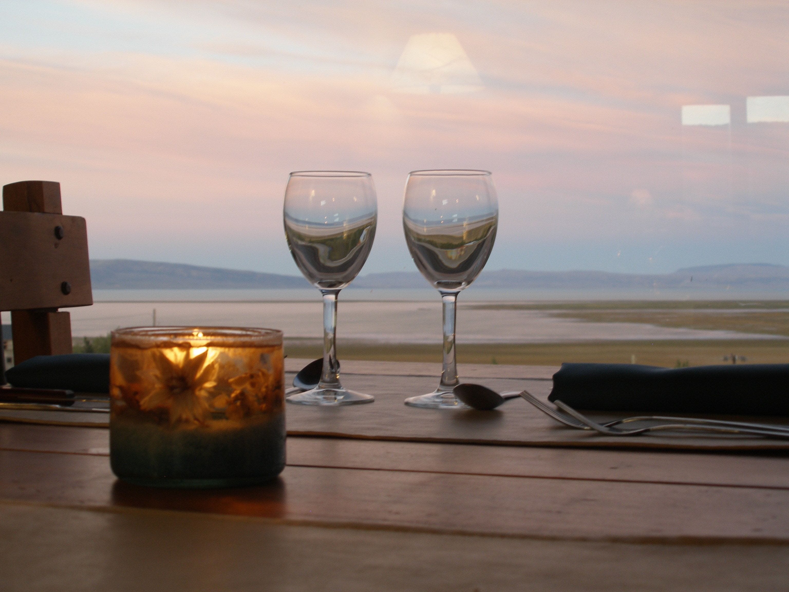 Two wine glasses on a wooden table with a candle, overlooking a body of water and distant mountains.