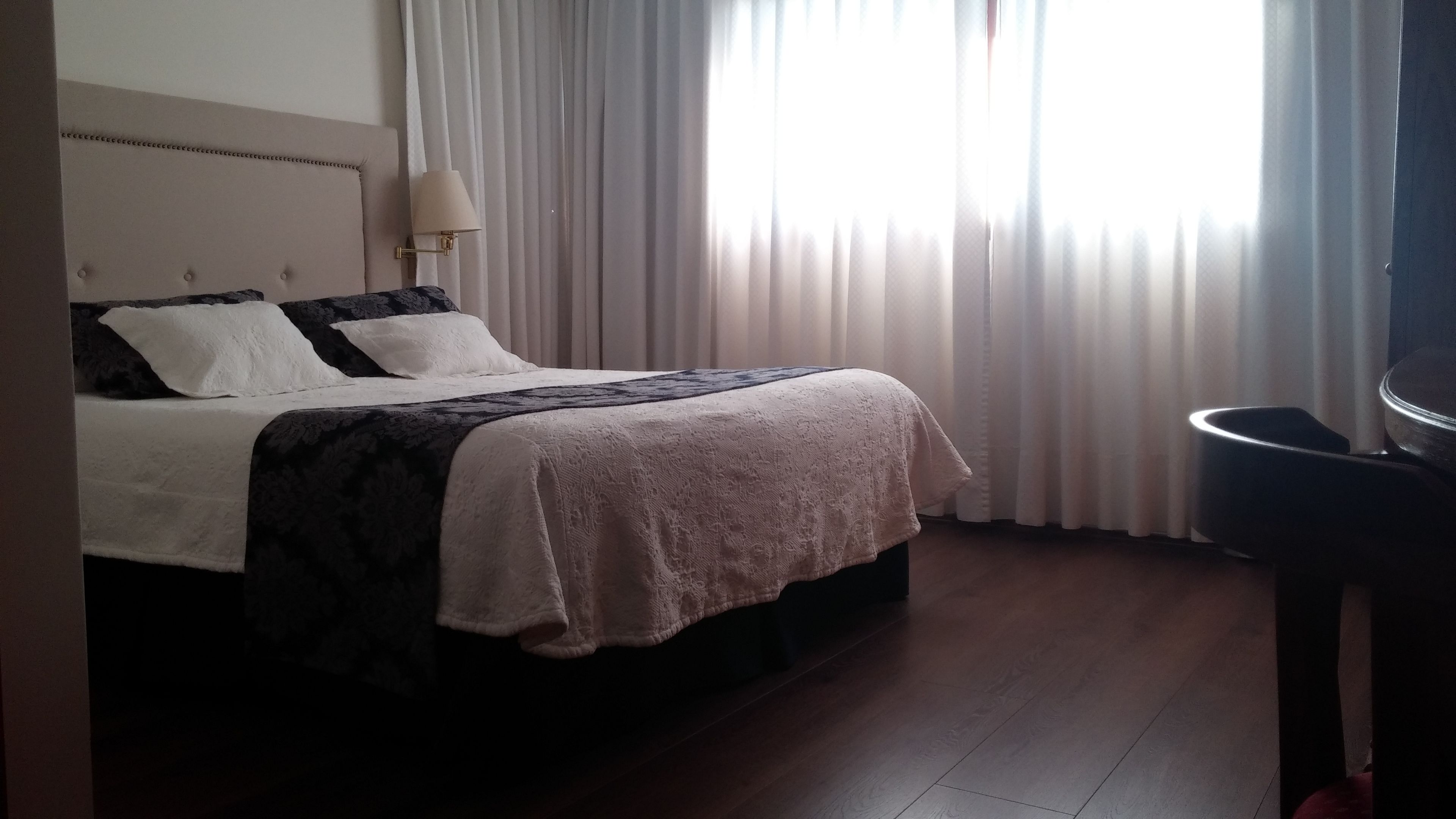 Habitación de hotel con cama, lámpara de noche y vistas al exterior a través de cortinas translúcidas.