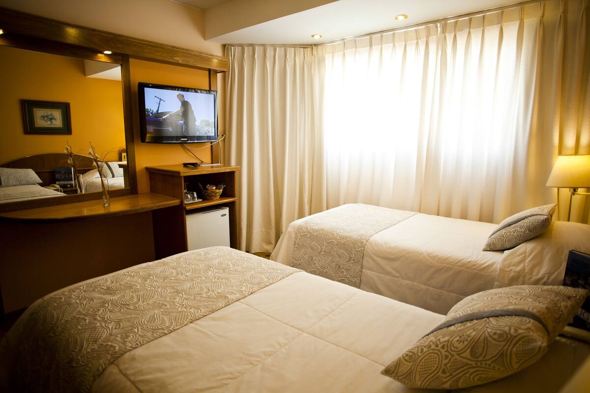 Quarto de hotel com duas camas, uma mesa de trabalho com televisão e uma janela com cortinas.