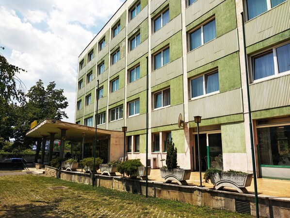 A multi-story building with a green facade, multiple windows, and a covered entrance area with potted plants.