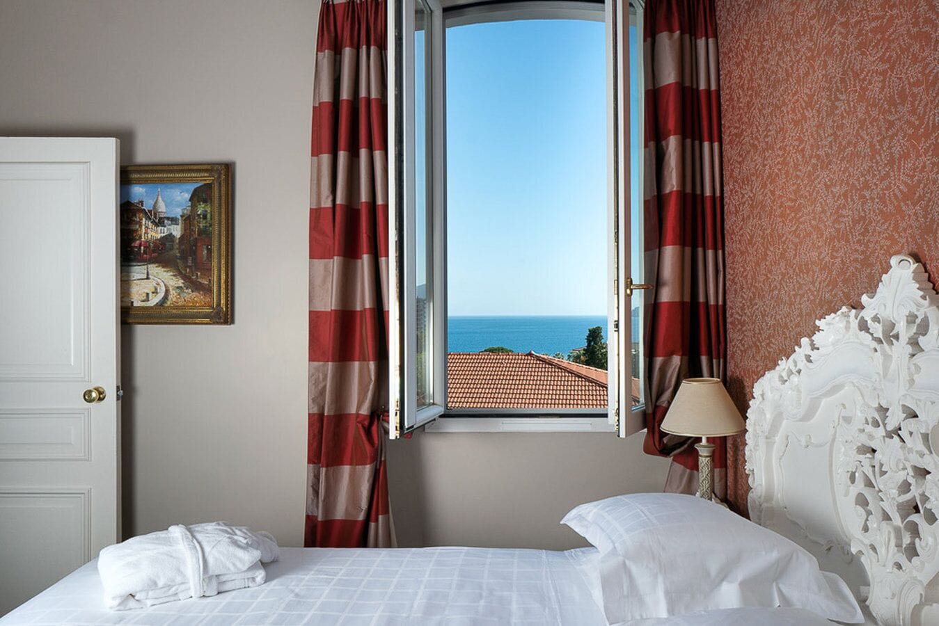 Une chambre d’hôtel avec un lit, un tableau, une fenêtre avec des rideaux et une vue sur la mer.