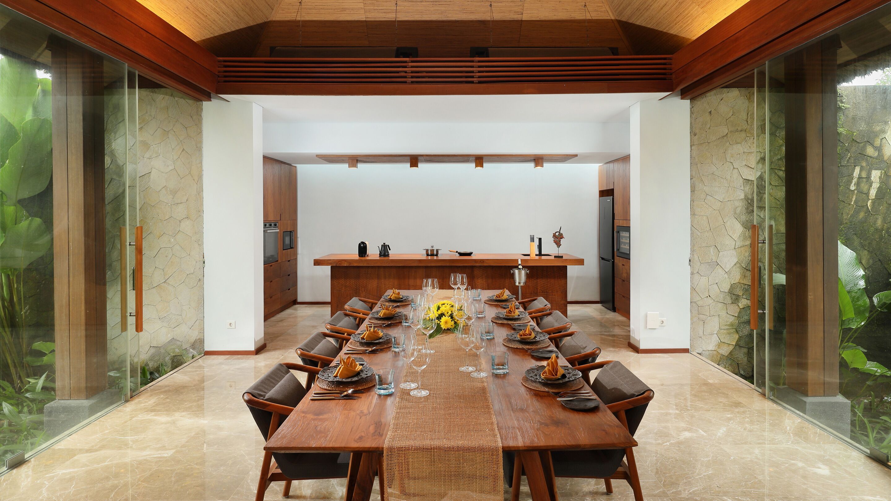 A modern dining area with a long wooden table set for a meal, flanked by glass walls and a kitchen in the background.