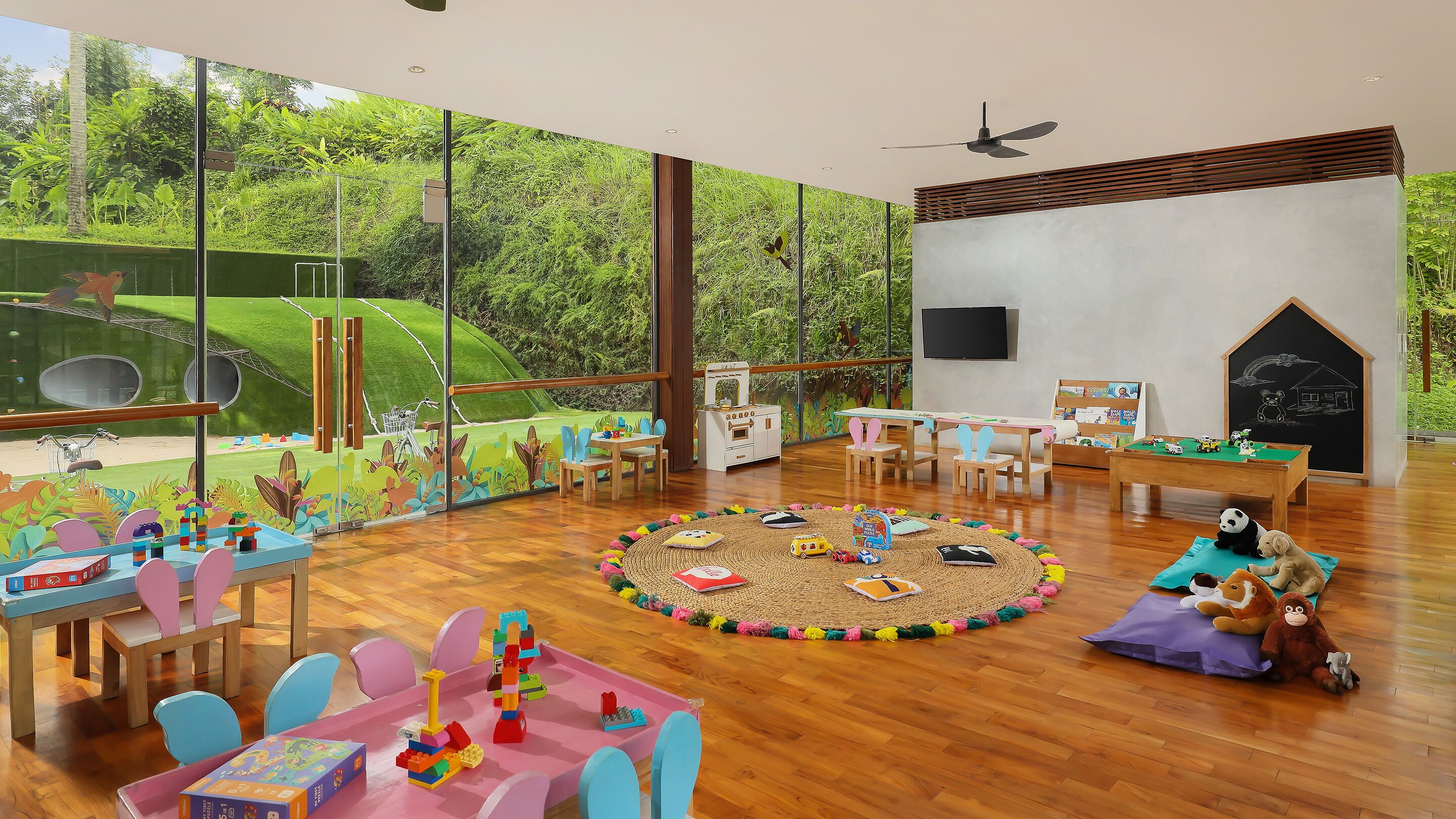 A spacious room with large windows, a wooden floor, a ceiling fan, and a children's play area with toys and a chalkboard.