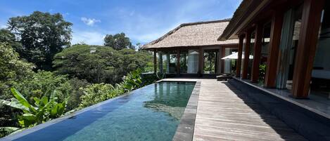 A clear pool with a wooden deck, surrounded by lush greenery and a modern house with large windows.