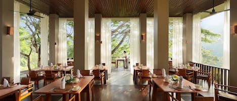 A restaurant with wooden tables and chairs, large windows with curtains, and a view of greenery.