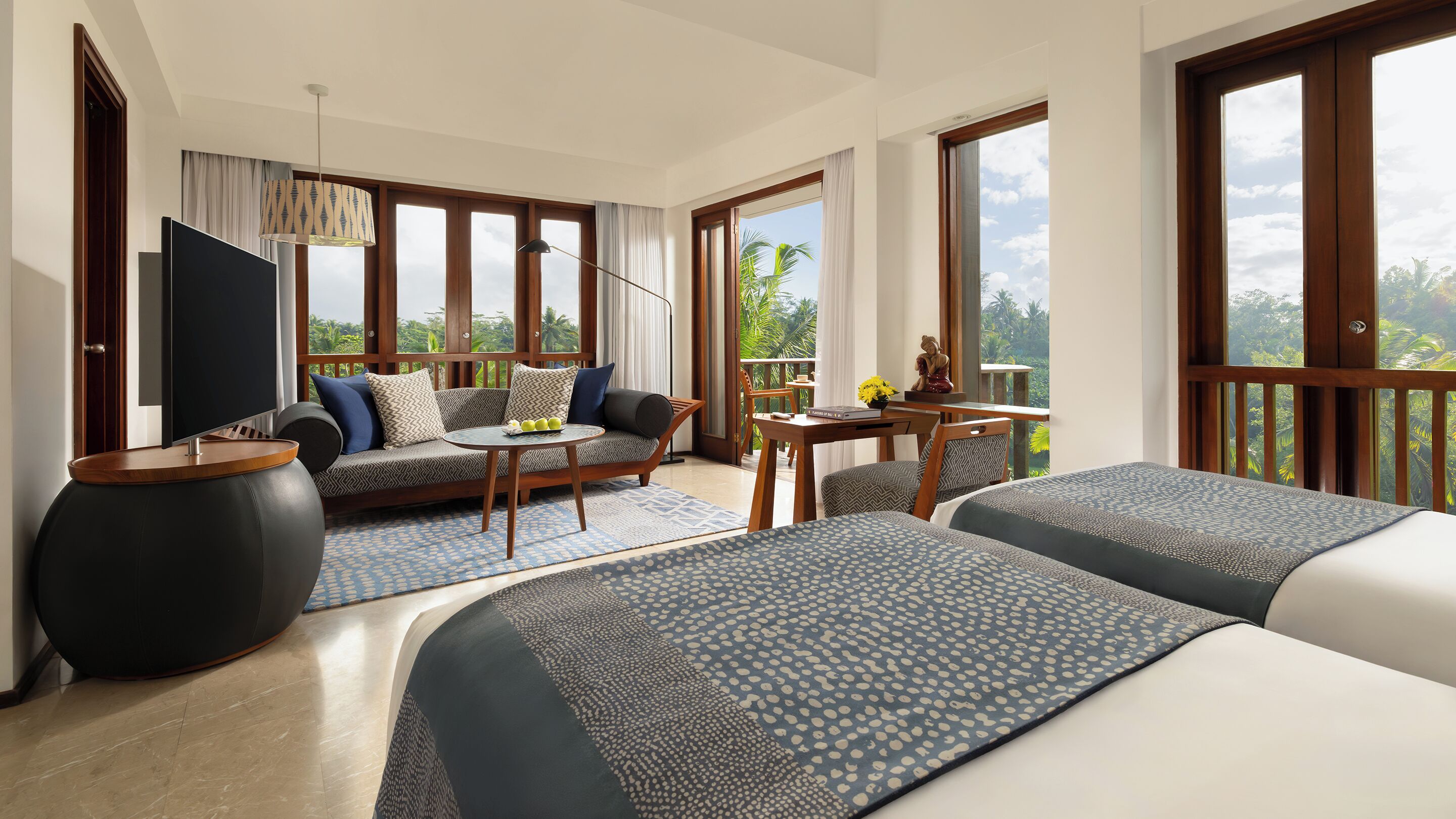 Habitación de hotel con sofá, dos camas, una mesa de comedor y ventanas amplias con vistas a zonas verdes.