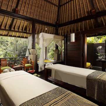 A room with two beds, a wooden table, chairs, and a view of lush greenery.