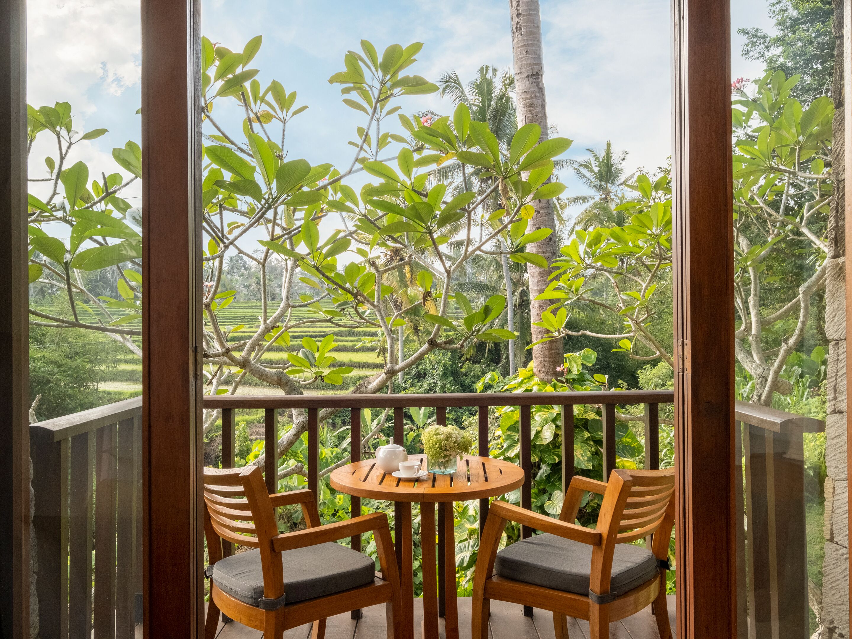 A wooden balcony with two chairs and a table, overlooking a lush garden with various plants and a palm tree.