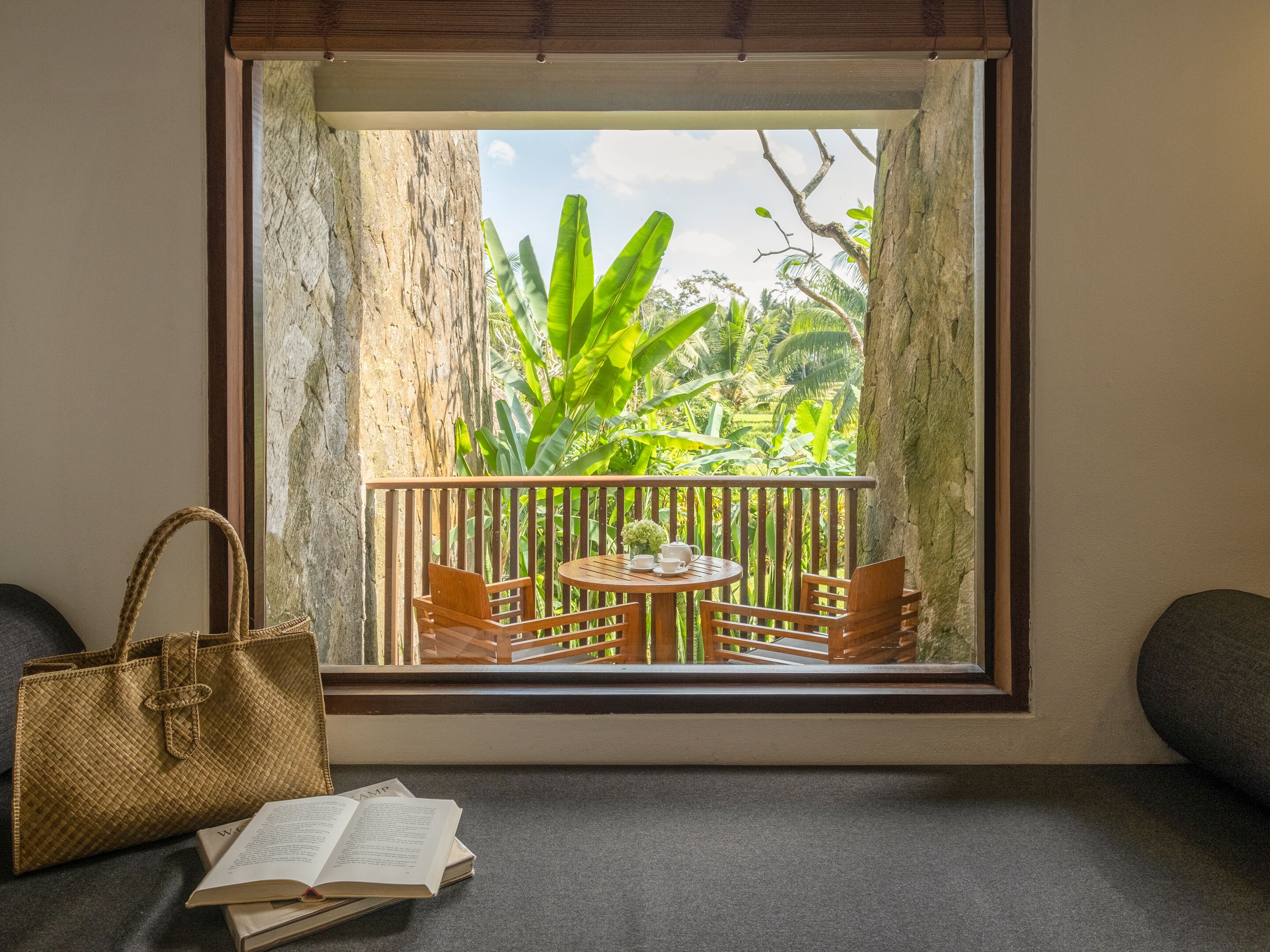 A balcony with a wooden table and chairs, a view of lush greenery, and a woven bag on the floor.