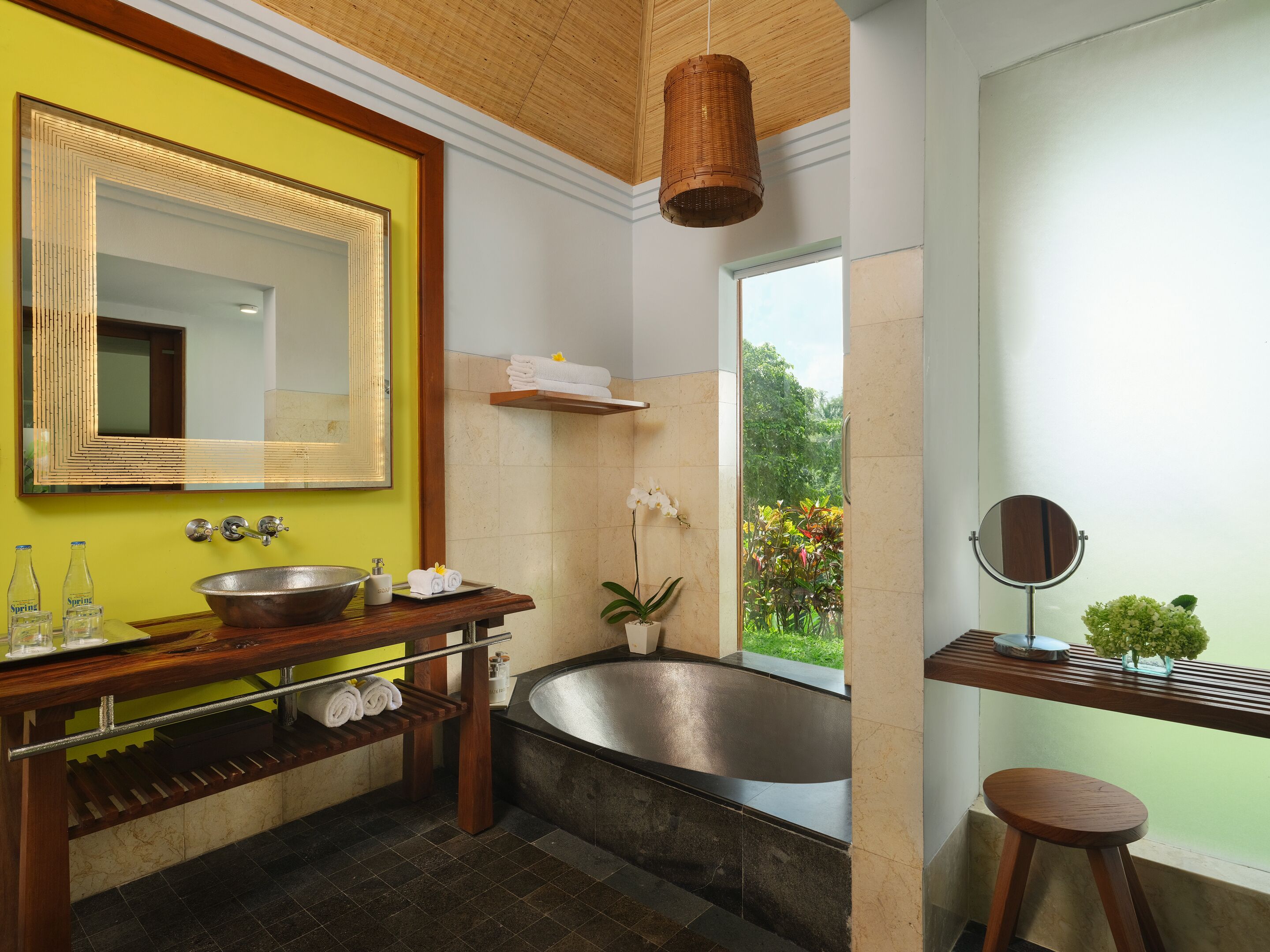 A modern bathroom with a large mirror, a bathtub, and a window with a view of greenery.