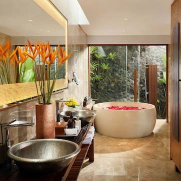 A modern bathroom with a large bathtub, a wooden vanity, a large mirror, and a view of a stone wall and greenery outside.