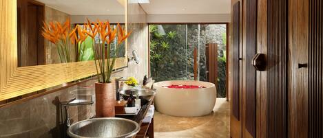 A modern bathroom with a large bathtub, a wooden vanity, a large mirror, and a view of a stone wall and greenery outside.