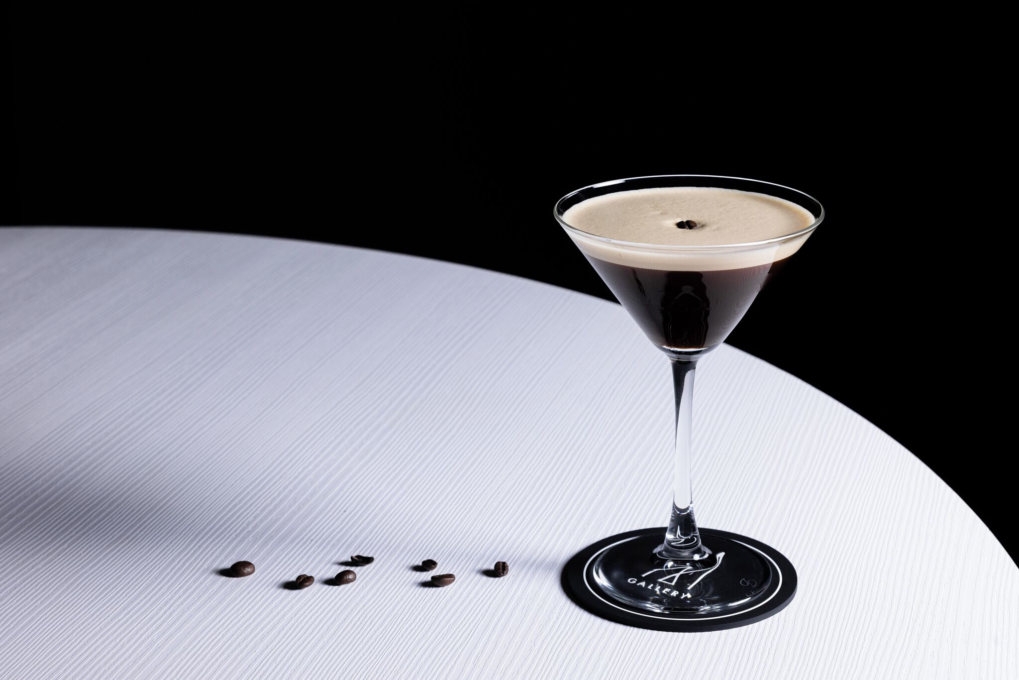 A martini glass with a dark liquid and a light foam on a white surface with scattered coffee beans.