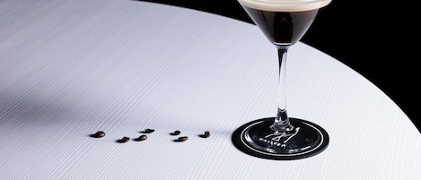 A martini glass with a dark liquid and a light foam on a white surface with scattered coffee beans.