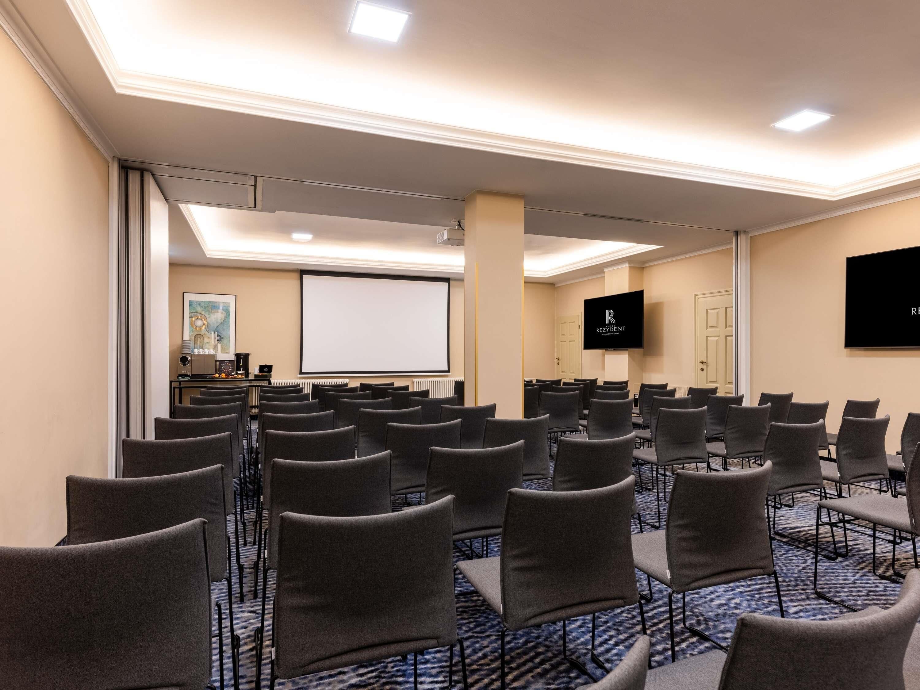 A conference room with rows of chairs, a projector screen, a television, and a door.
