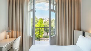 Hotellihuone, jossa on sänky, työpöytä ja näkymä parvekkeelle, jolta avautuu vihreää kasvillisuutta ja rakennuksia.