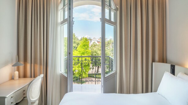 A hotel room with a bed, a desk, and a view of a balcony with greenery and buildings. - Rezydent Hotel Sopot - MGallery Collection (Sopot)