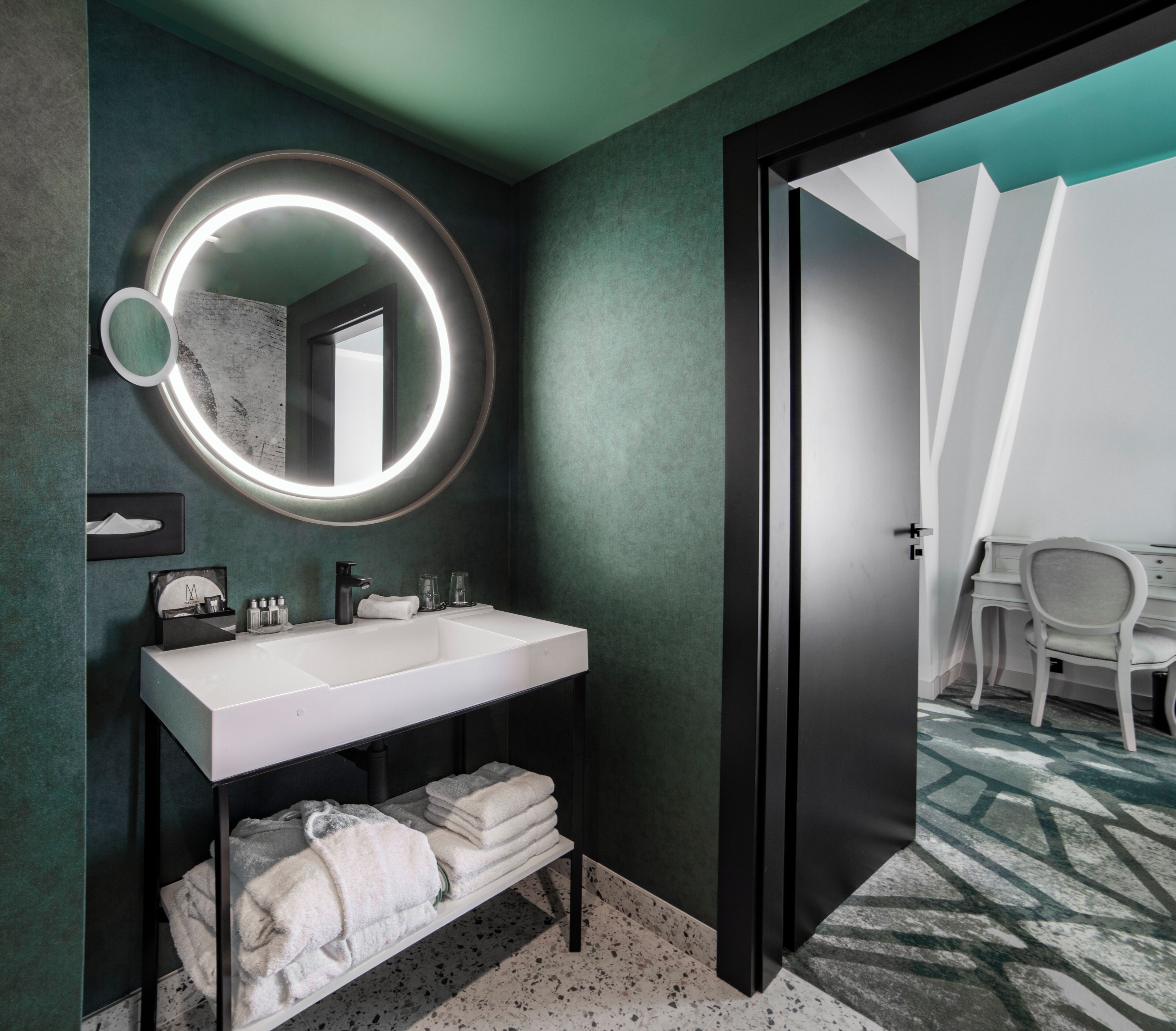 A modern bathroom with a white sink, a round mirror with a light, and a door leading to a bedroom.