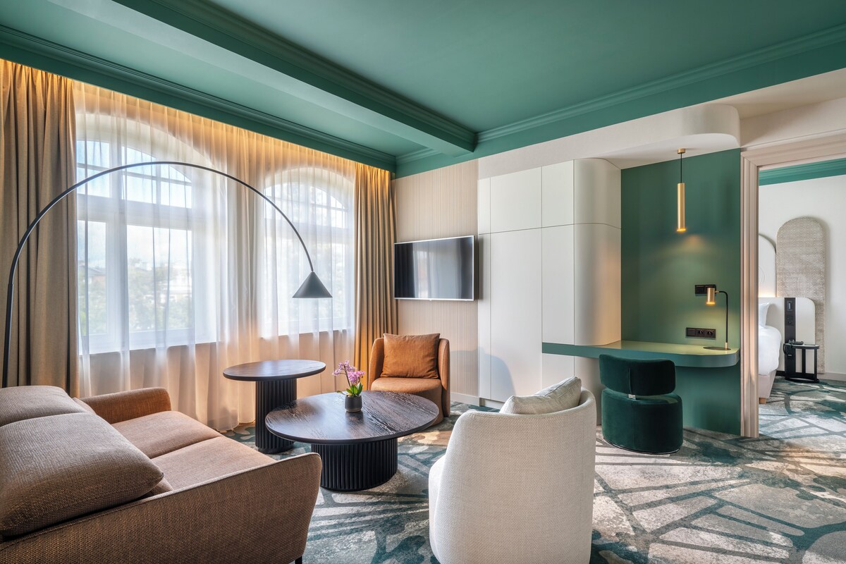 A modern hotel room with a sofa, armchair, round coffee table, and a small side table. There is a TV mounted on the wall, a desk with a lamp, and a patterned carpet.