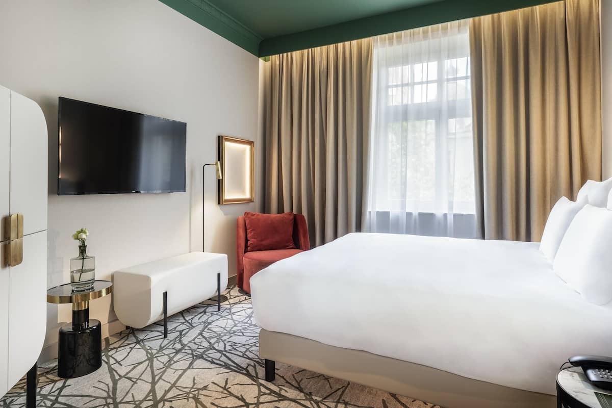 A hotel room with a large bed, a flat-screen TV, a bedside table with a lamp, a red chair, and a window with curtains.