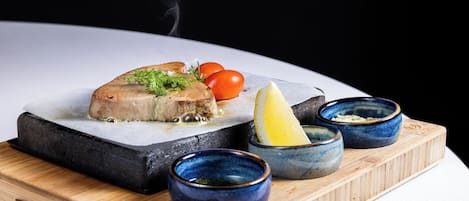 Un plato de pescado sobre una losa de piedra, con tomates cherry y gajos de limón, acompañado de recipientes pequeños con salsas sobre una tabla de madera.