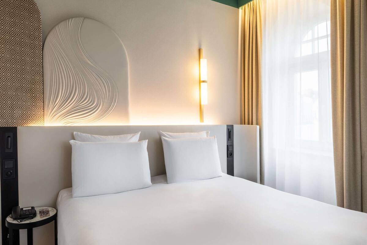 Ein modernes Hotelzimmer mit einem großen Bett, einem Nachttisch mit Telefon und Zeitschrift, einer Wandlampe und einem mit Vorhängen versehenen Fenster.