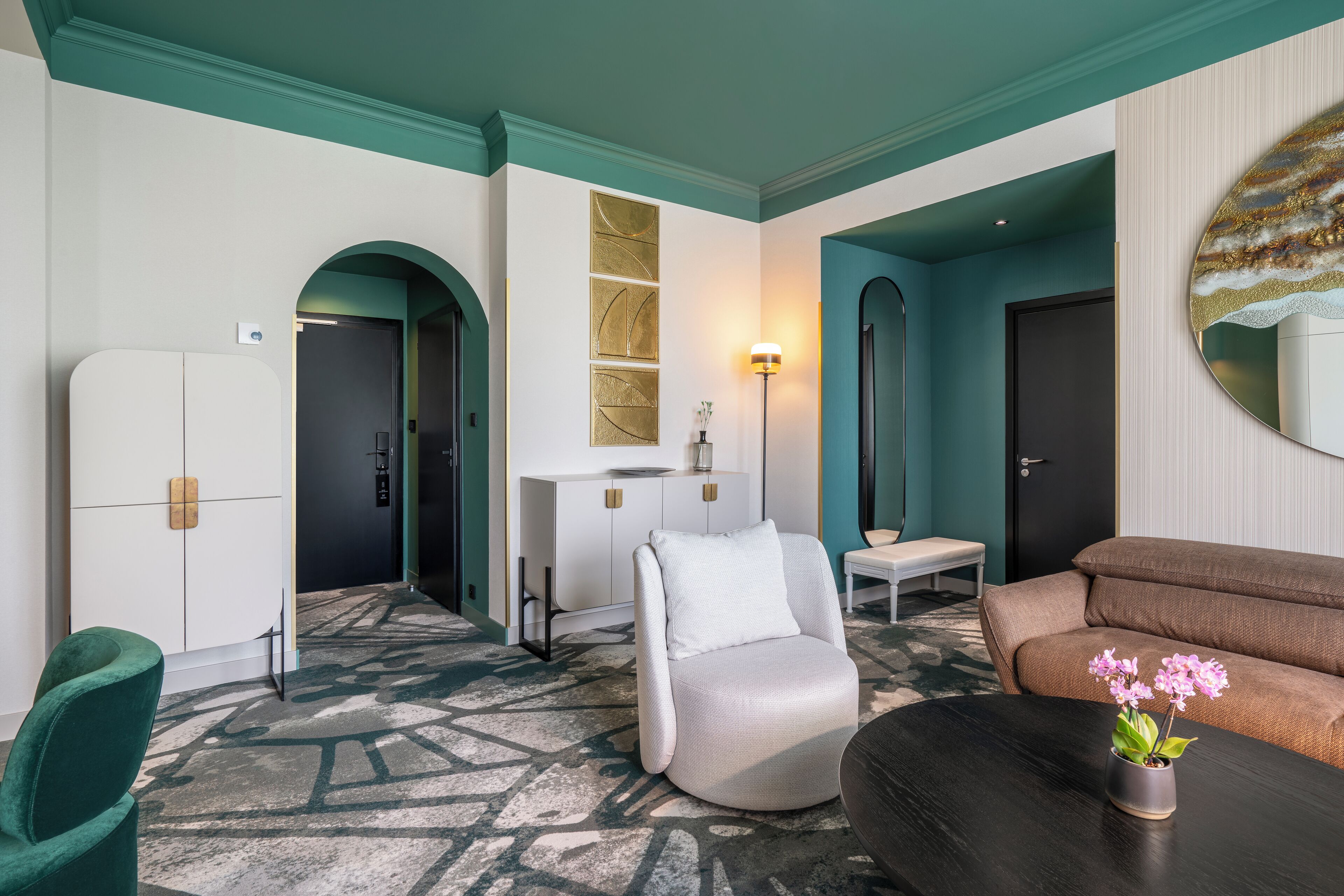 Et moderne hotelværelse med en teal farvet accentvæg, et hvidt skab, en grøn lænestol, en brun sofa og et lille bord med en vase blomster.