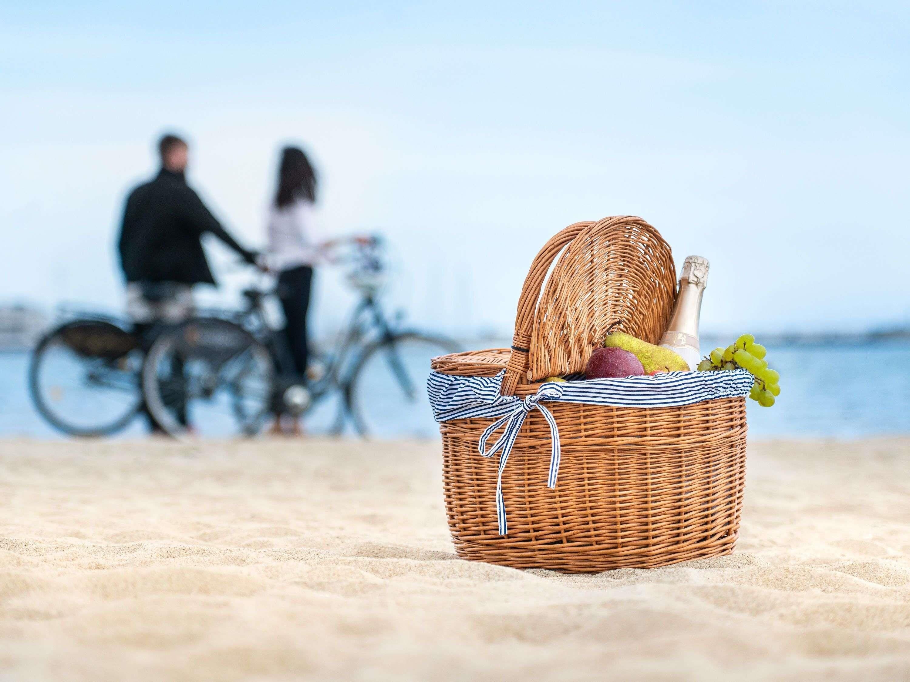 Ein aus Weidwerk geflochtener Picknickkorb mit Speisen und Getränken an einem sandigen Strand, im Hintergrund sind Fahrräder zu sehen.