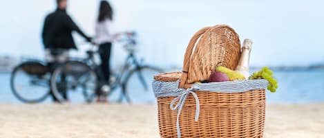 Una cesta de mimbre con comida y bebidas en una playa arenosa, con bicicletas al fondo.