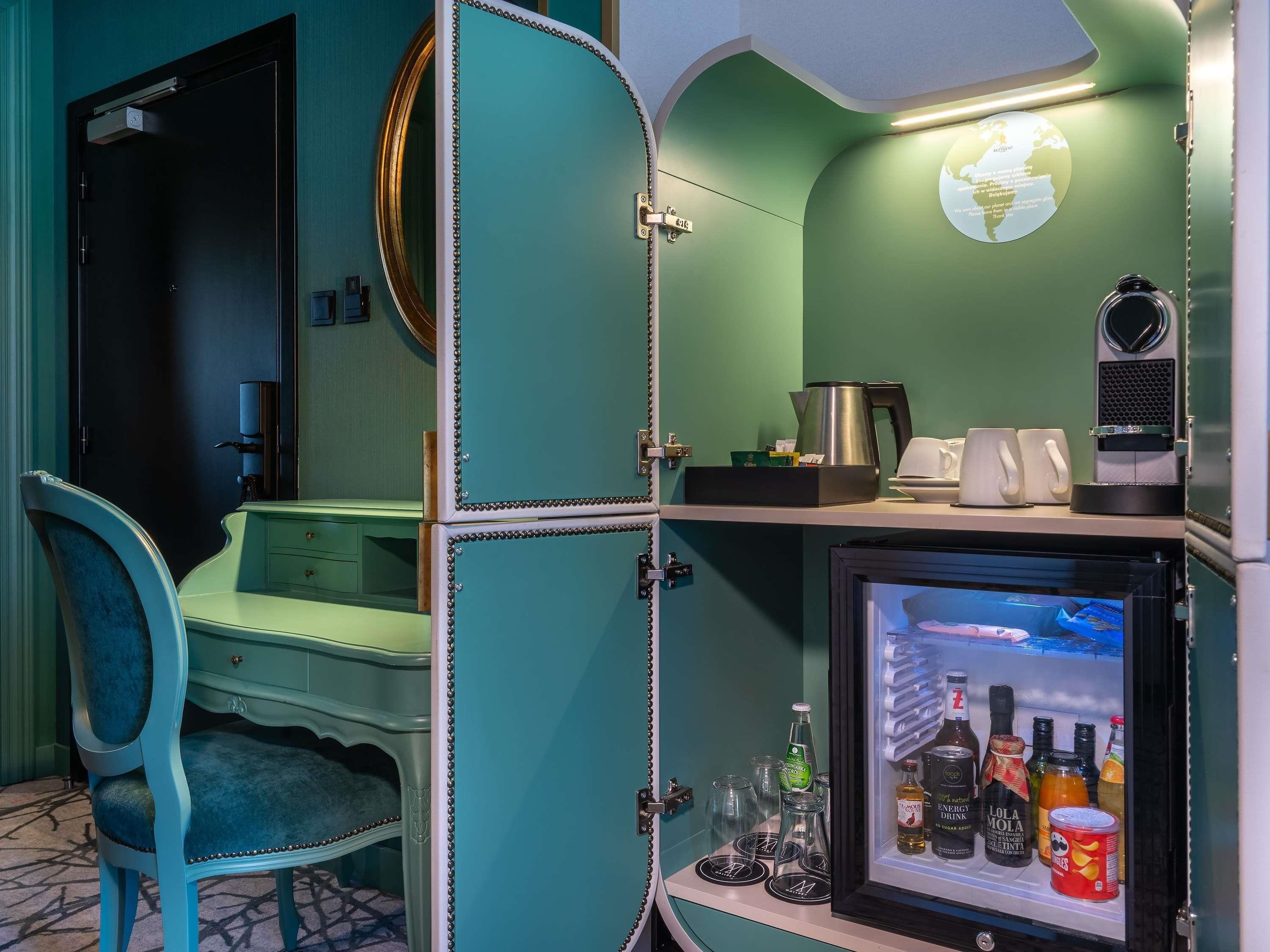 Une pièce aux murs de couleur bleu-vert, avec un lit, un bureau et un réfrigérateur rempli de boissons.