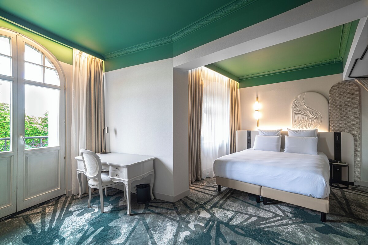 A hotel room with a bed, desk, and chair, large windows with curtains, and a patterned carpet.