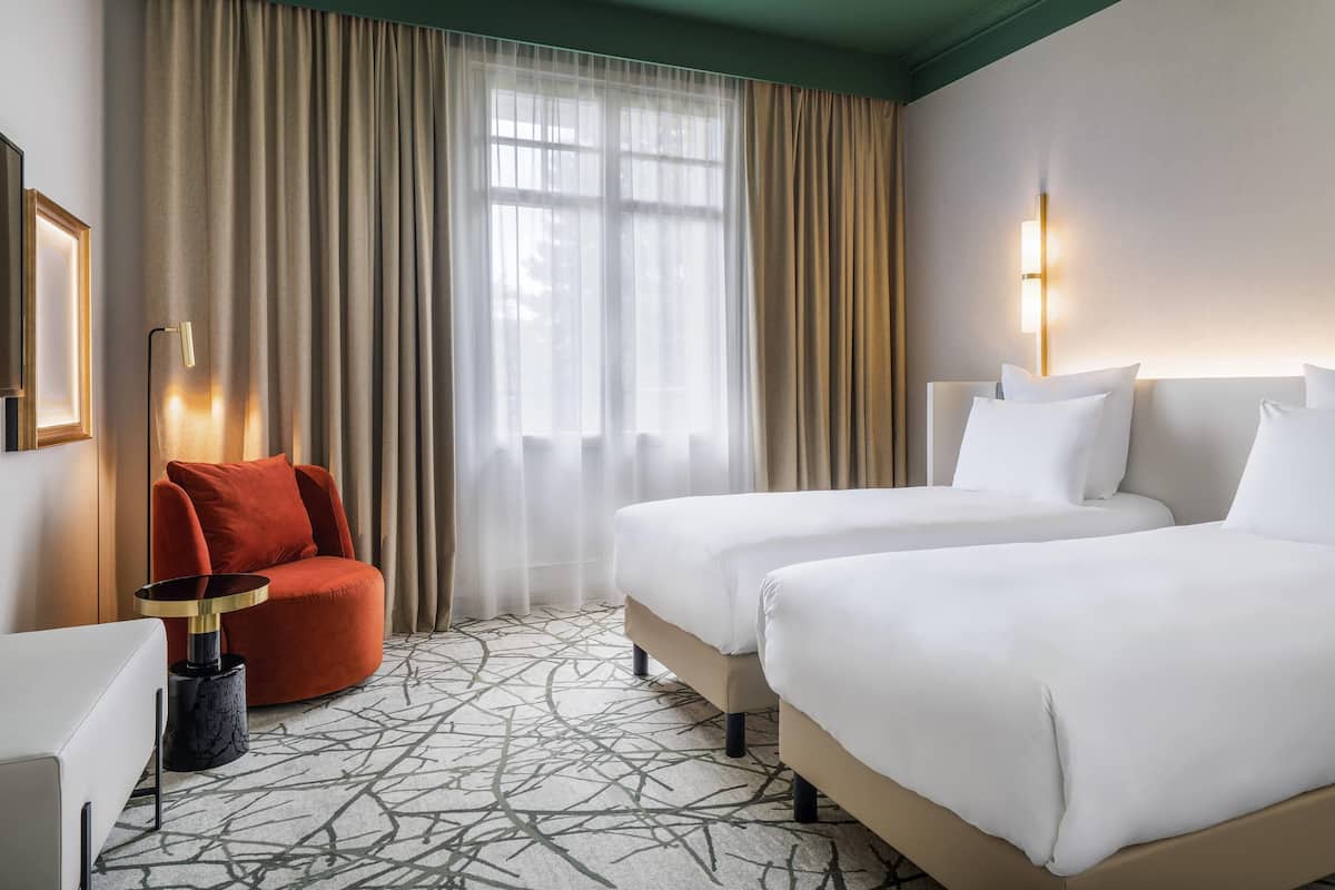 A hotel room with two beds, a red armchair, a small table, and a large window with sheer curtains.