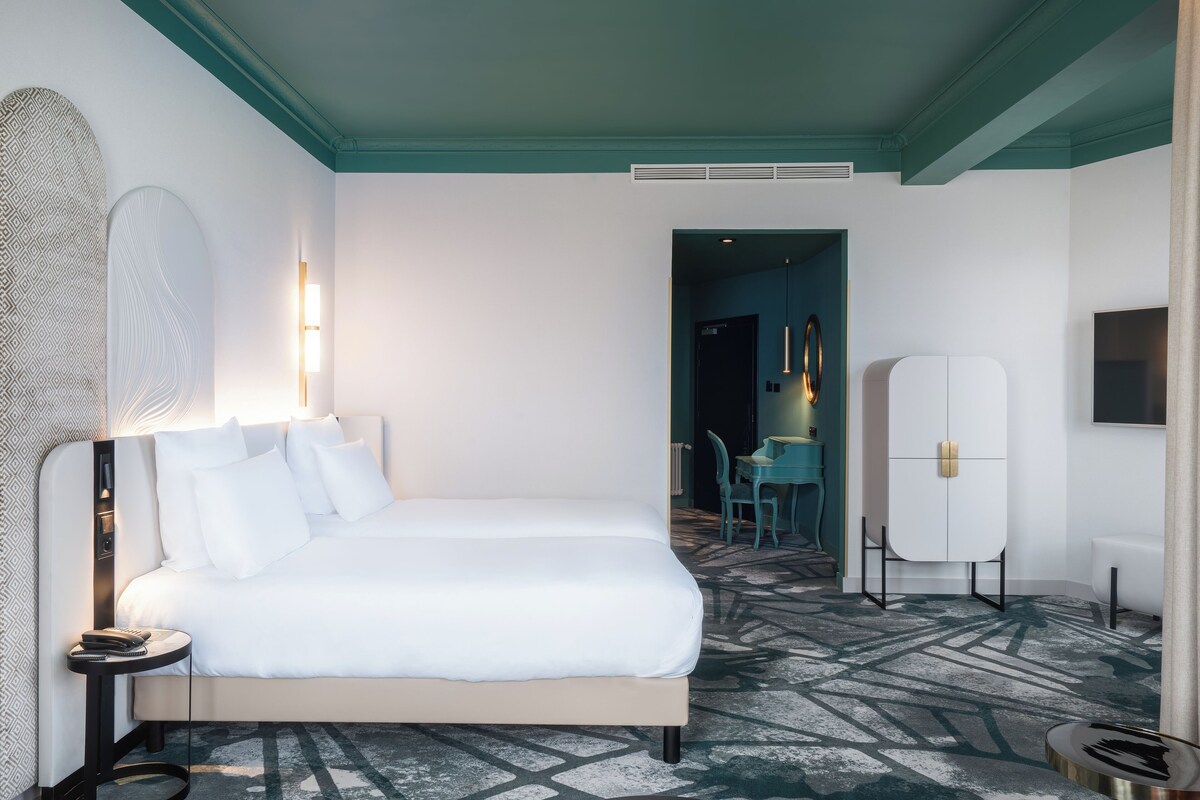 A modern hotel room with a large bed, a bedside table, a wall-mounted TV, and a view into the dining area.
