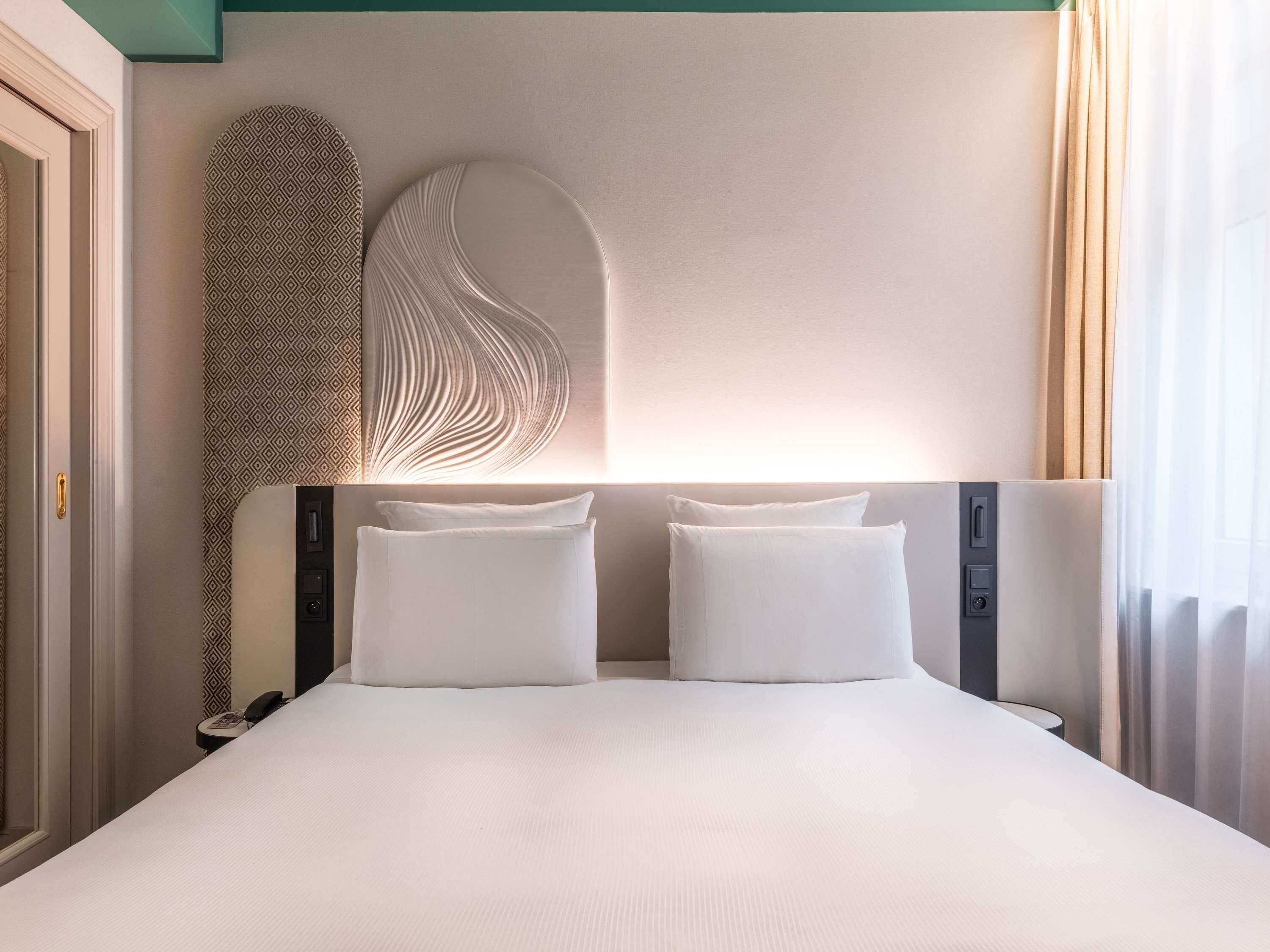 A hotel room with a large bed, two pillows, a patterned wall, and a decorative wall piece.