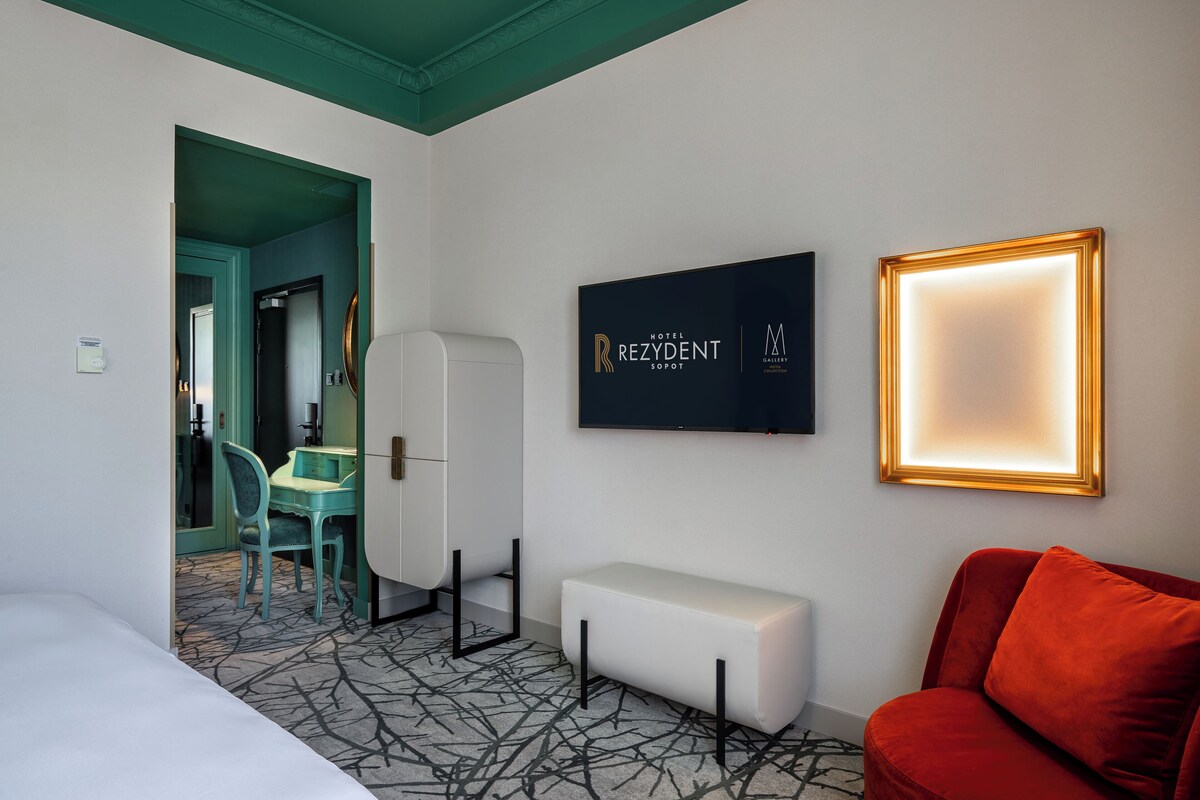 A hotel room with a bed, a desk with a chair, a bench, and a red sofa. There is a framed picture on the wall and a sign that reads "Hotel Rezydent Sopot."