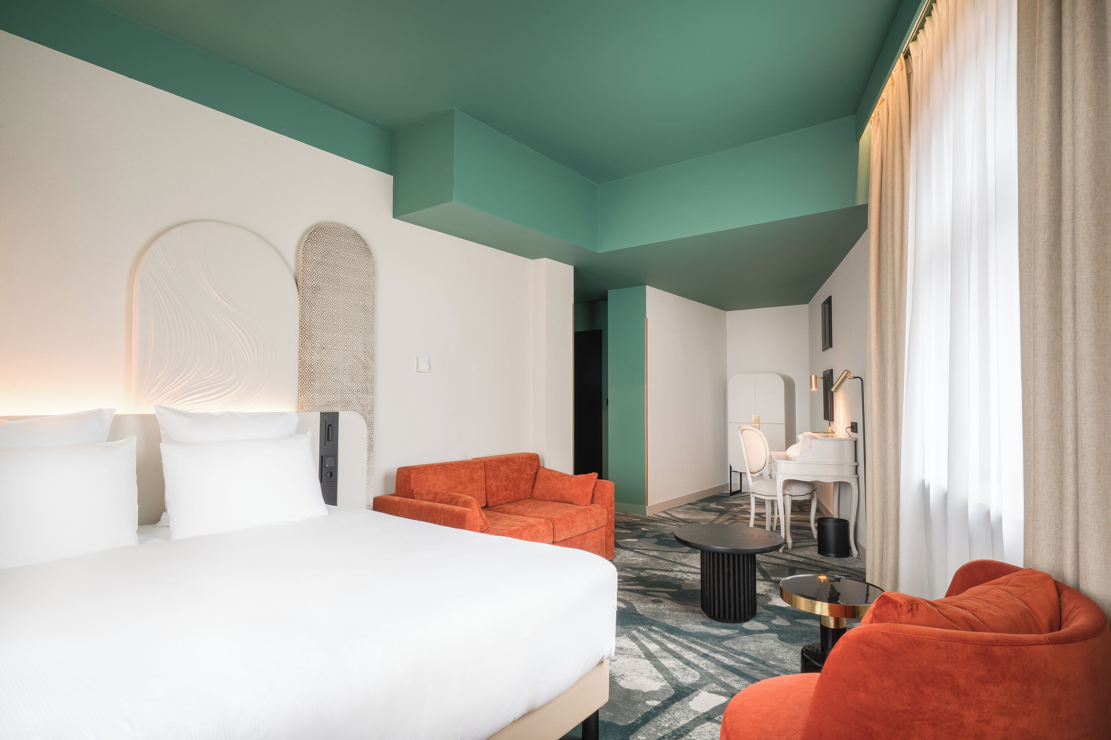 A hotel room with a bed, orange armchairs, a small table, and a piano in the background.