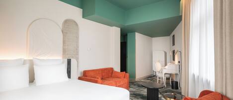 Habitación de hotel con cama, sillones naranja, una pequeña mesa y un piano al fondo.