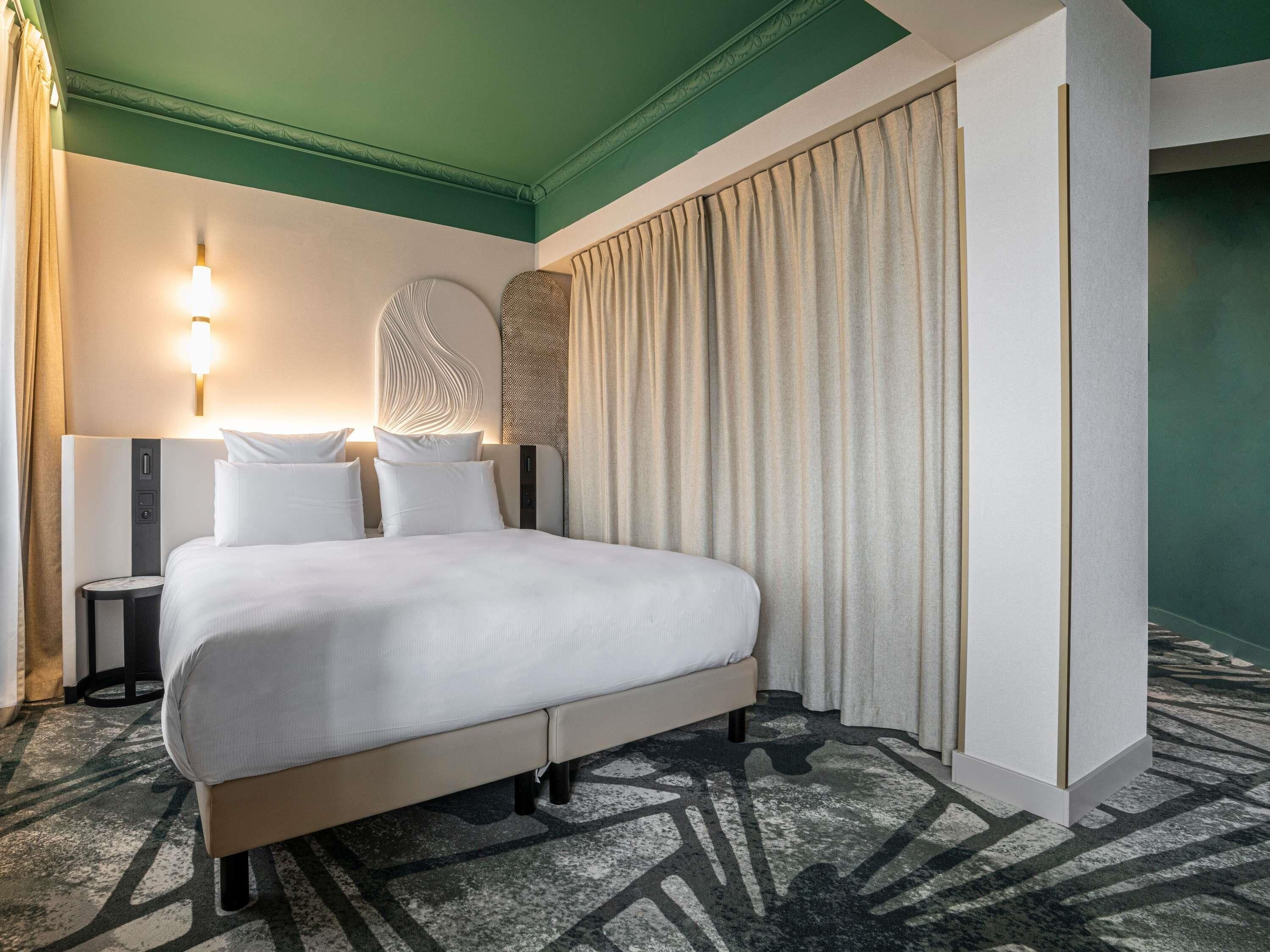 A hotel room with a bed, a nightstand, a wall-mounted lamp, and a large window with curtains.