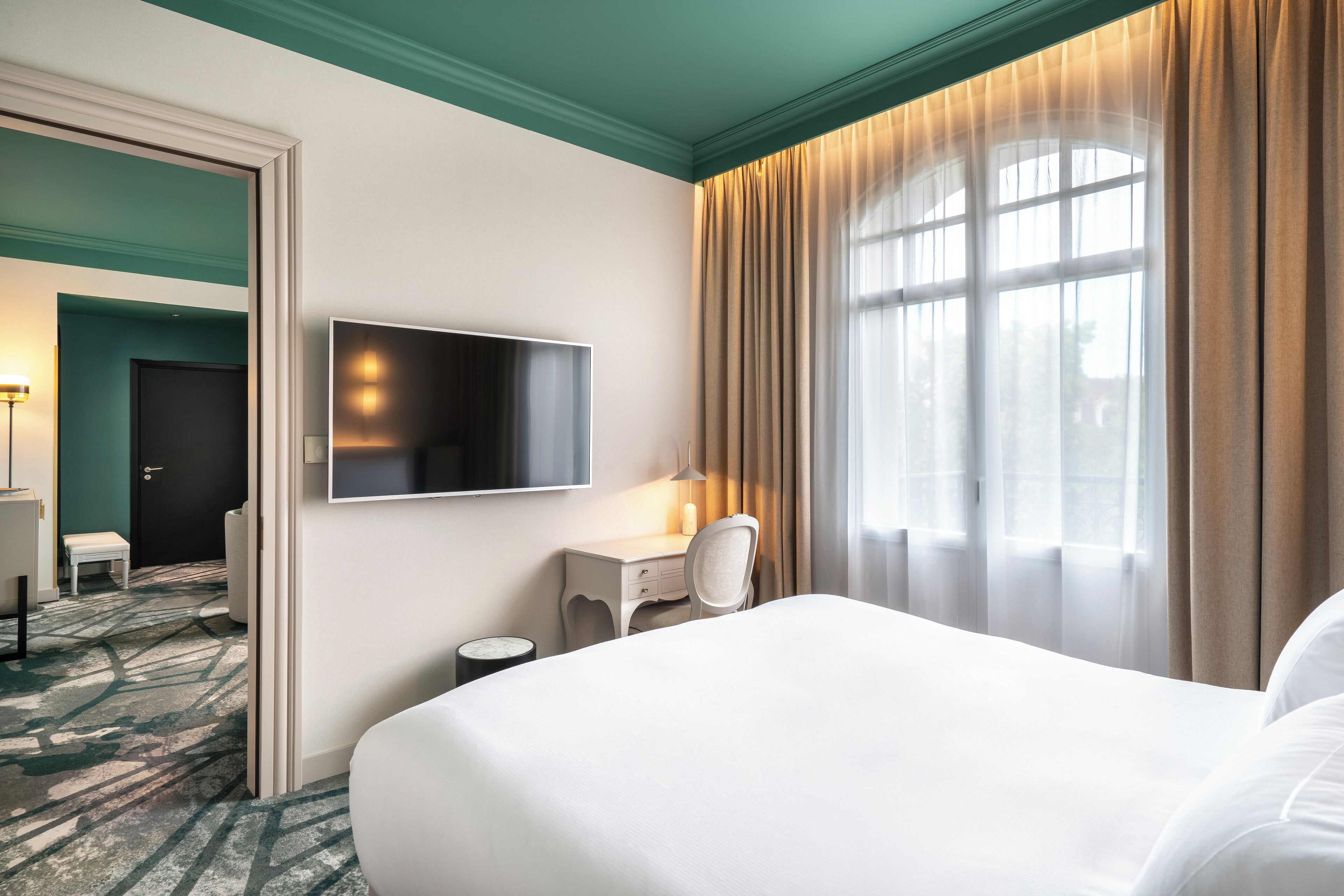 Ein Hotelzimmer mit einem großen Bett, einem Schreibtisch mit Stuhl, einem Flachbildfernseher und einem Fenster mit Vorhängen.