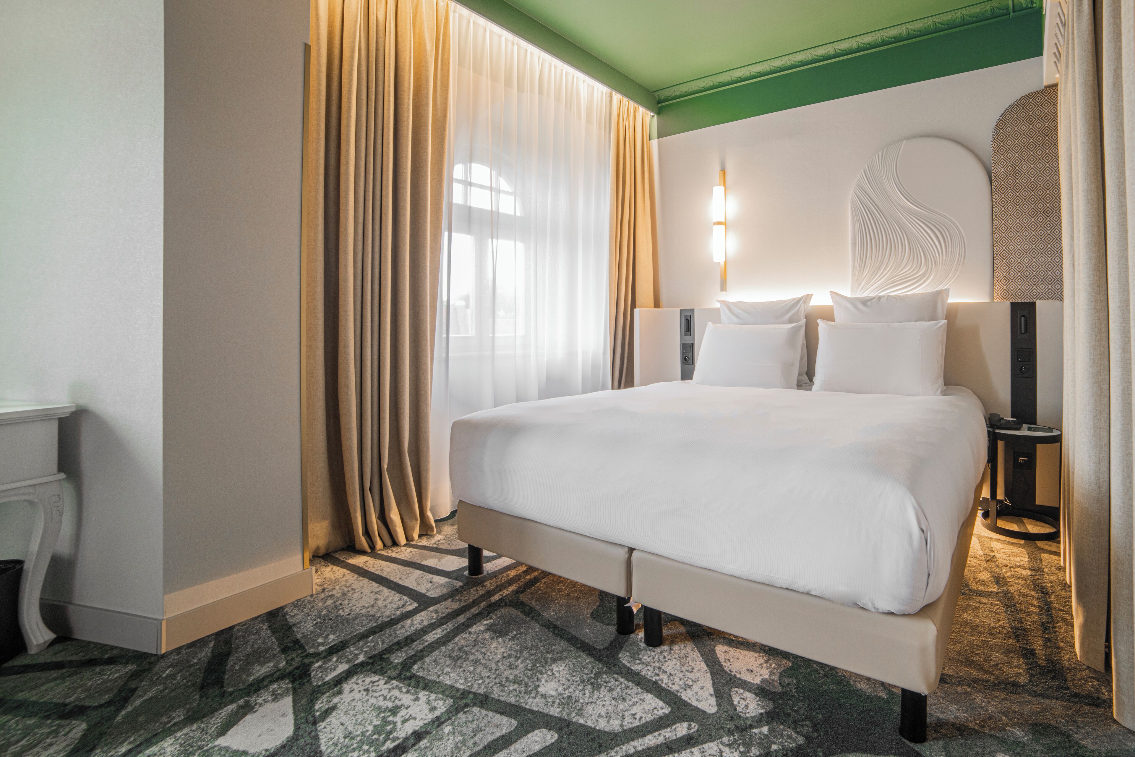 Une chambre d’hôtel avec un grand lit, une table de chevet, une commode, une lampe fixée au mur et un élément décoratif mural.